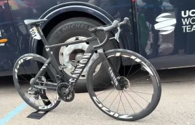 Una bicicleta de ruta Canyon negra con calcomanías blancas y un juego de ruedas Zipp 2026 está estacionada frente a un autobús del equipo mundial UCI, con su etiqueta con número visible en el cuadro, lista para rodar más rápido en el pelotón.