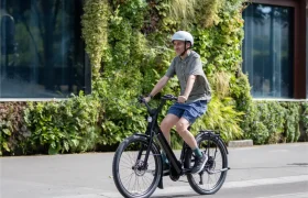 Una persona con casco circula en una bicicleta eléctrica «Moustache Lundi 27» por una calle de la ciudad, con un jardín vertical verde como telón de fondo.