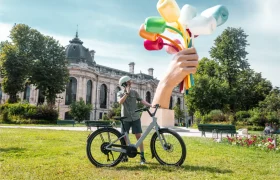 Una persona está de pie junto a una bicicleta «Moustache Lundi 27» sobre la hierba, hablando por teléfono, cerca de una escultura de una mano gigante que sostiene objetos de colores, con un edificio histórico al fondo, lo que transmite el animado ambiente de la ciudad.