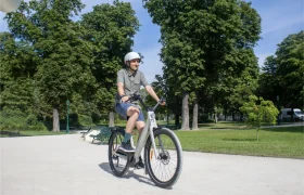 Una persona con casco va en una bicicleta eléctrica «Moustache Lundi 27» por un sendero de un parque urbano, rodeada de árboles y césped verde en un día soleado.
