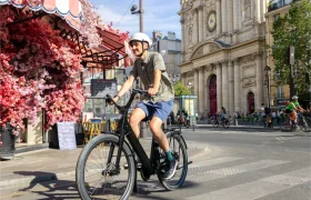 Una persona que lleva casco va en una bicicleta «Moustache Lundi 27» por una calle de la ciudad, cerca de un edificio con grandes columnas y una cafetería adornada con flores rosas.